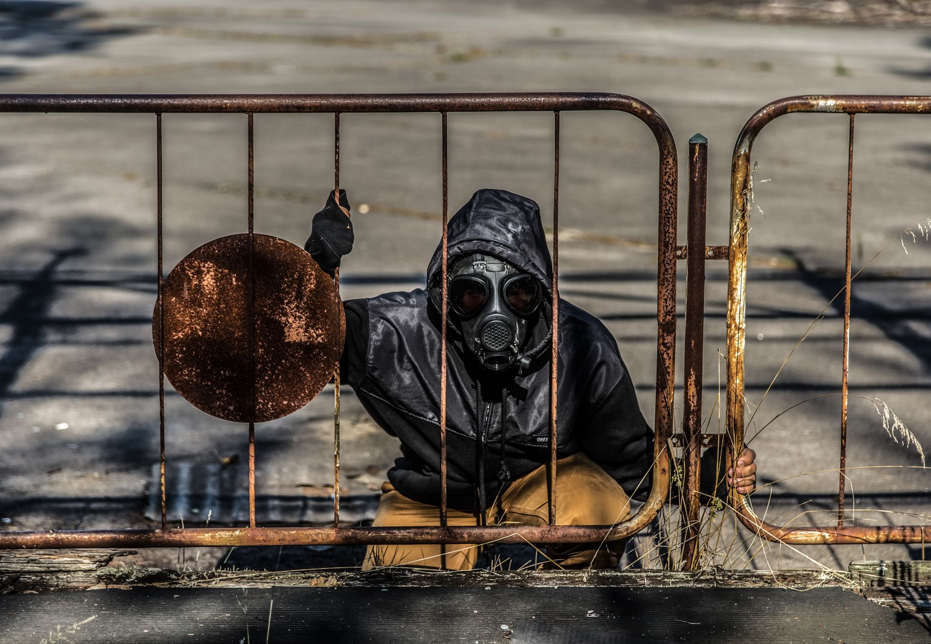 Person wearing a gas mask crouches behind a rusty fence, depicting a post-apocalyptic scene.