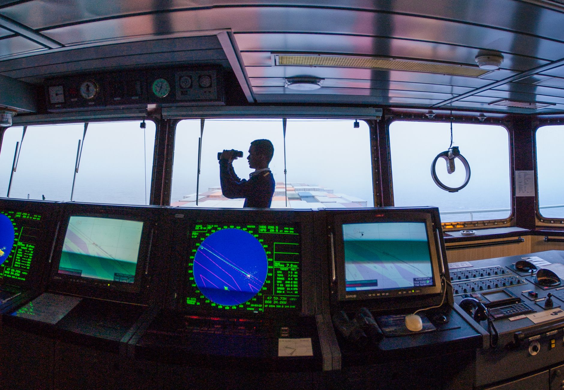 Silhouette of a navigator using binoculars on a cargo ship's bridge, surrounded by navigation equipment.