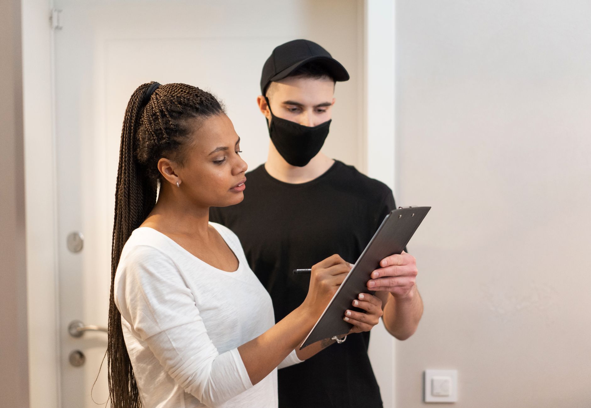 A woman signs a clipboard held by a delivery man wearing a face mask, illustrating a delivery service interaction.